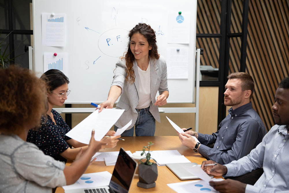 Smiling female company leader ceo give paper report business plan to diverse employees at group office meeting, businesswoman manager conducting team briefing work on paperwork at boardroom table