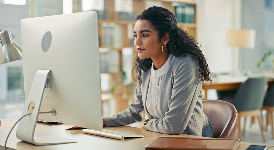 Focused businesswoman using a desktop computer, seated in a stylish office space with shelves.